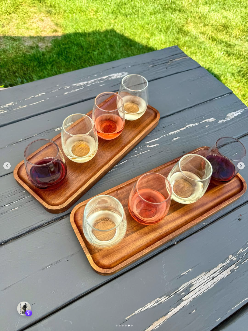 Two wooden wine flight trays with four glasses each — reds, rosés, whites — on a weathered grey picnic table with grass behind