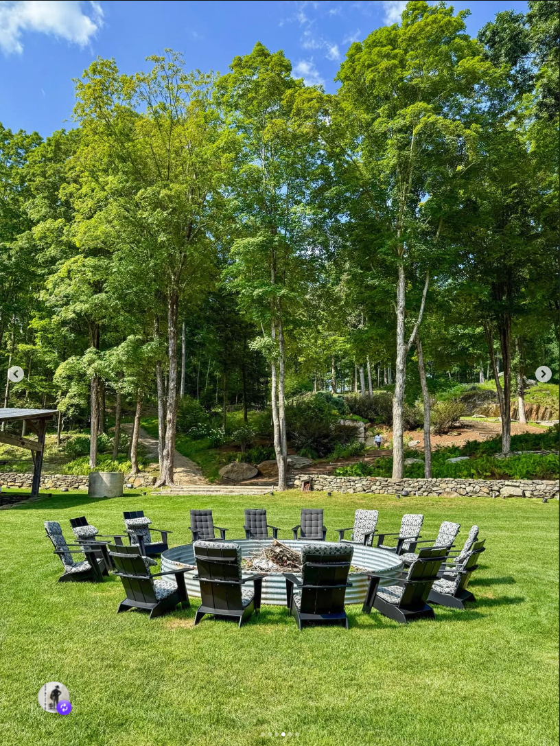 Cushioned Adirondack chairs in a circle around a steel firepit ring, deep green woods behind