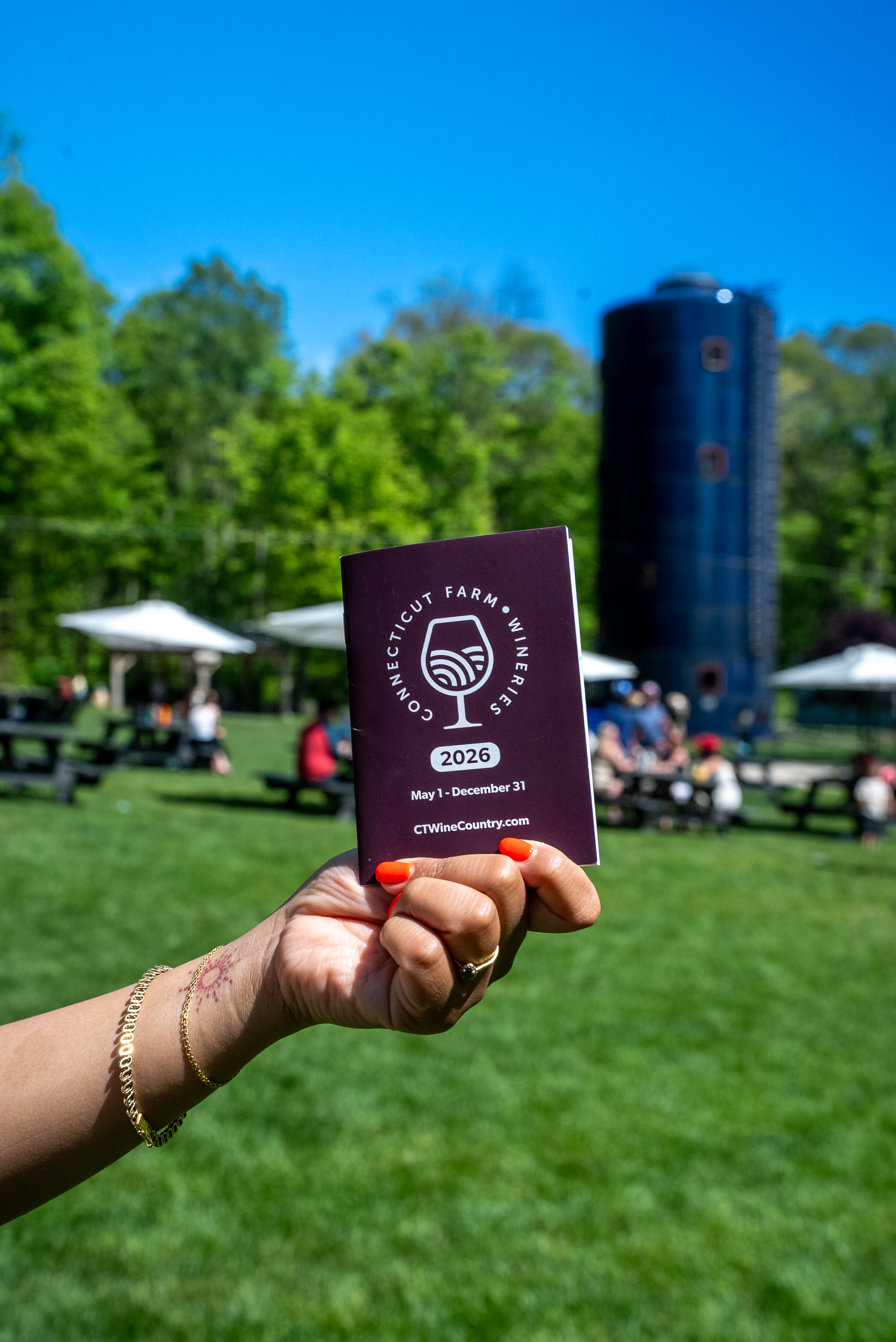 Hand holding a single closed passport up against the Spring Hill property — blue silo and white umbrellas behind