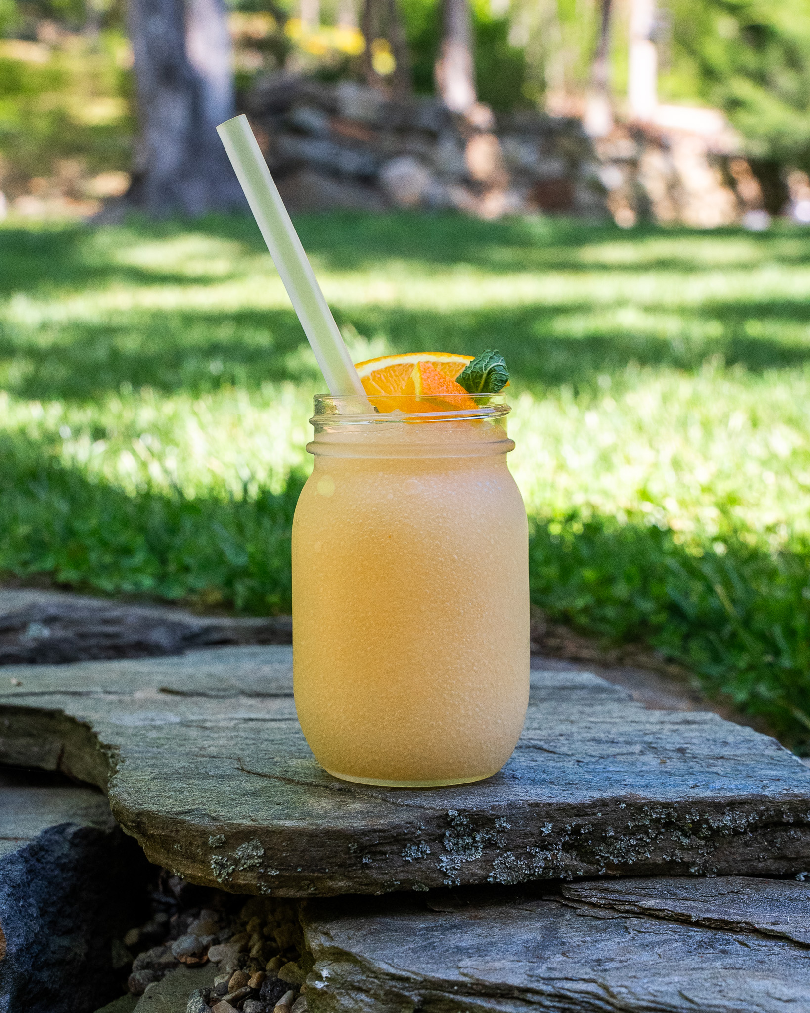 Frosted mason jar with pale orange Summer Breeze frozen cocktail, orange wheel and mint garnish, white straw, on a flat stone slab with lawn behind