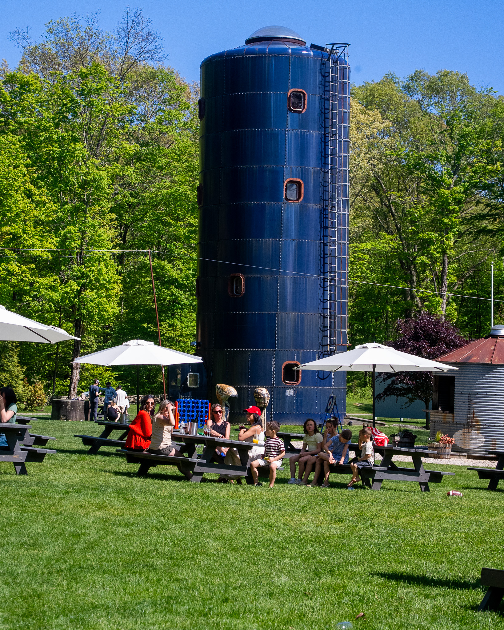 Wide shot of the blue silo with families seated at picnic tables under white umbrellas, Connect 4 game on the table, kids of all ages, deep green woods behind