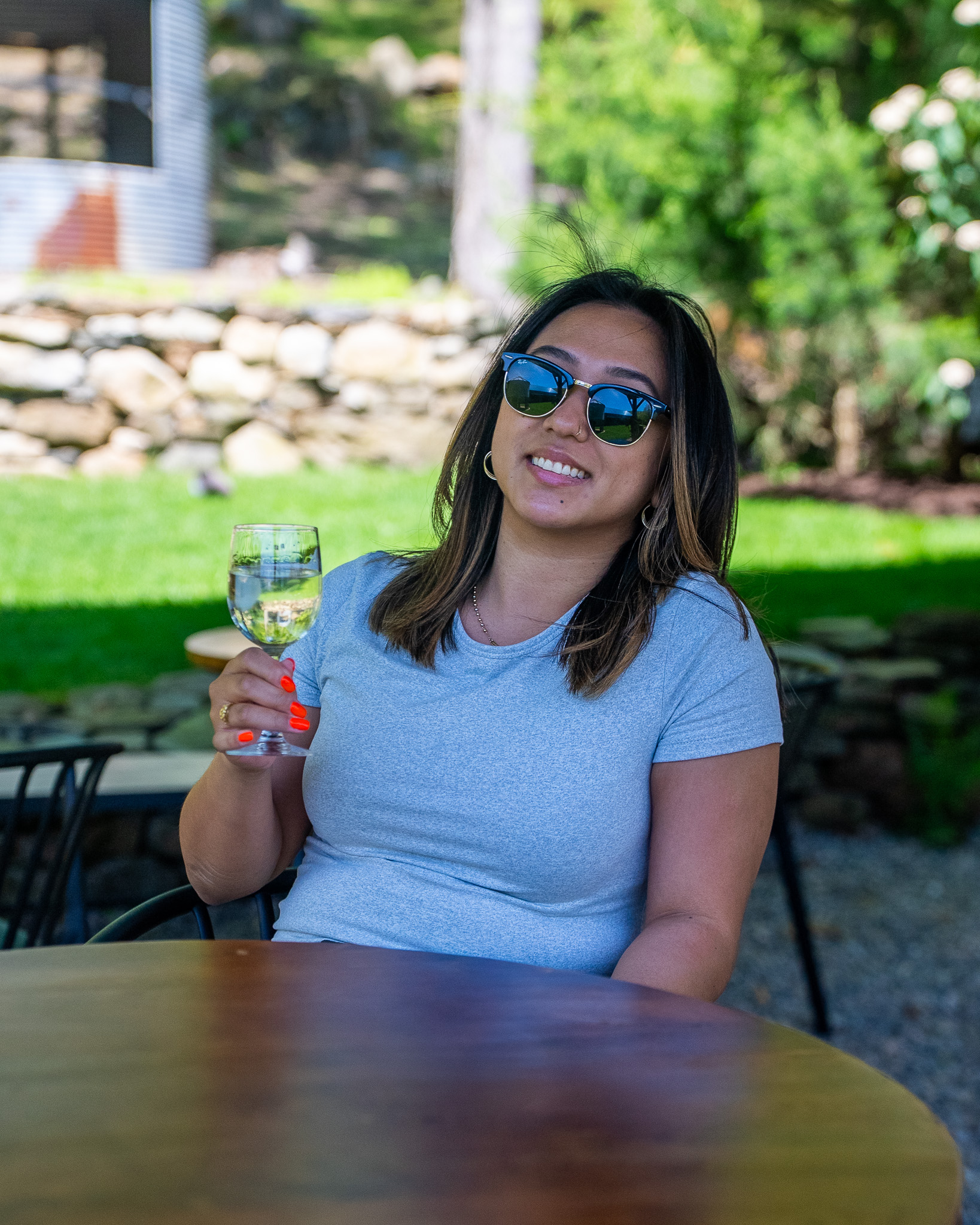 Young woman in her 20s smiling, sunglasses, grey tee, holding a glass of white wine at a circular cafe table with a stone wall and lawn behind
