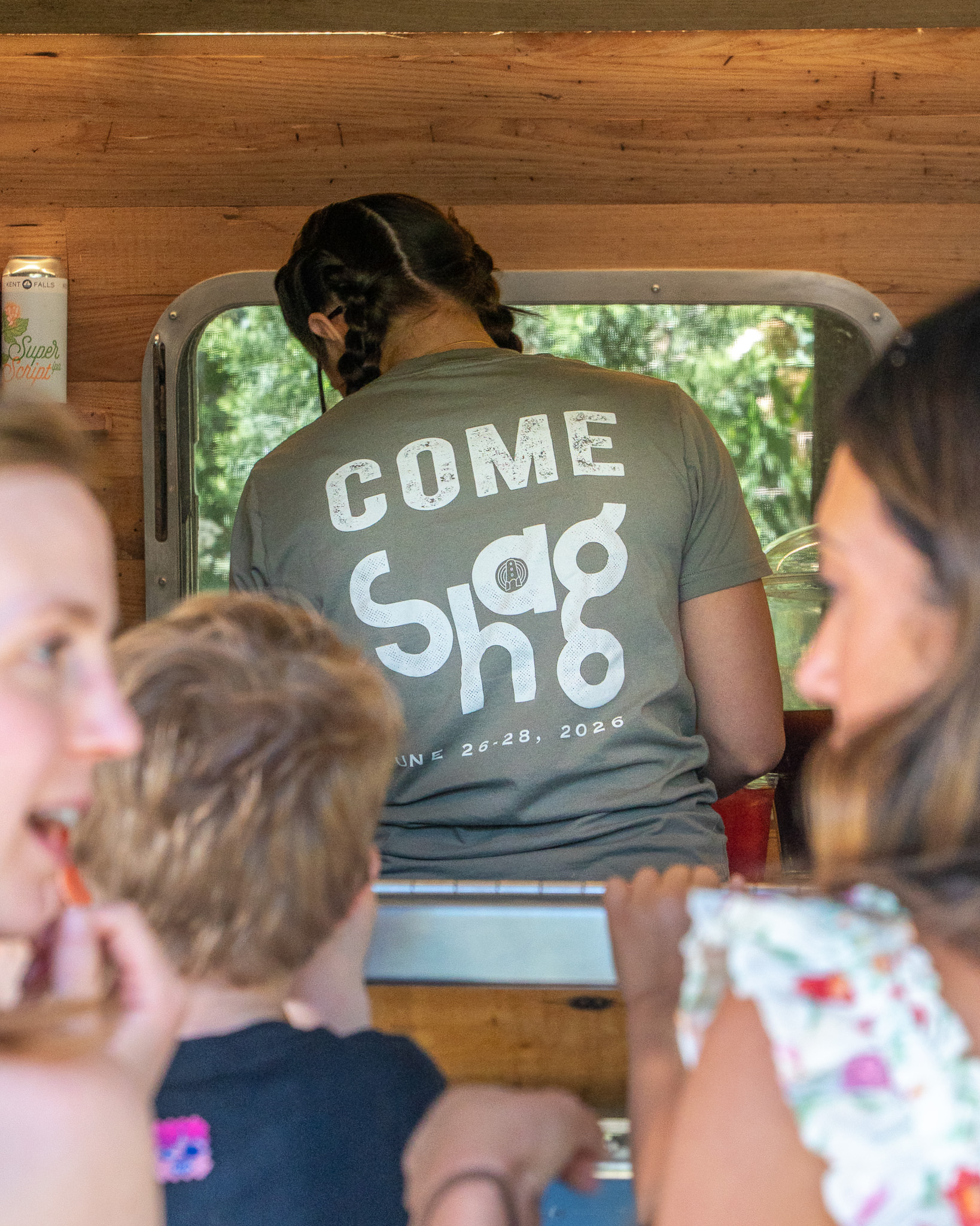 Staff member in olive-grey COME SHAG JUNE 26-28 2026 tee at the Airstream service window, two young guests in soft-focus foreground